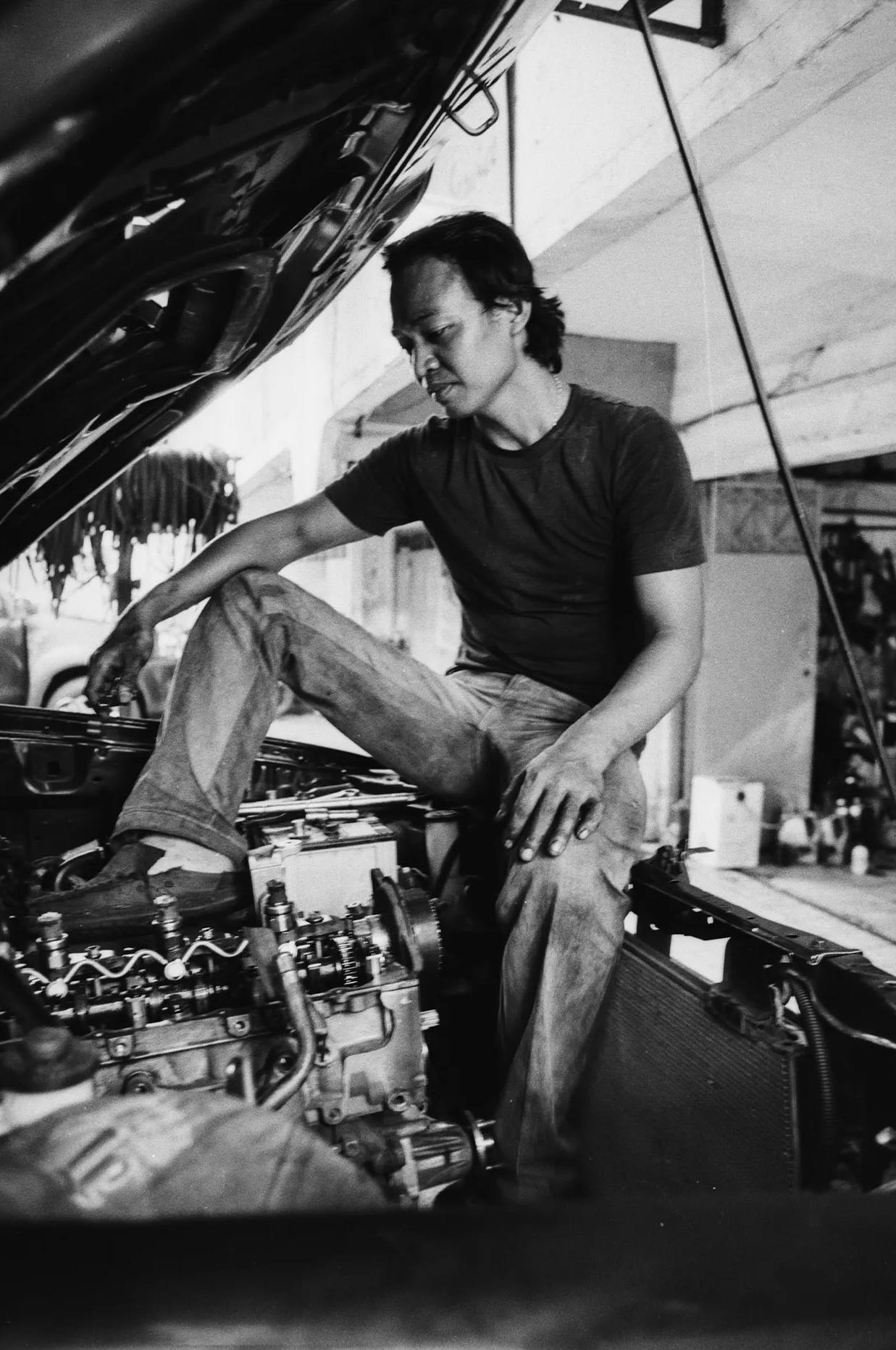 Uomo seduto nel vano motore di una vecchia auto americana in un’officina meccanica, ambientazione grezza e autentica, foto in bianco e nero.