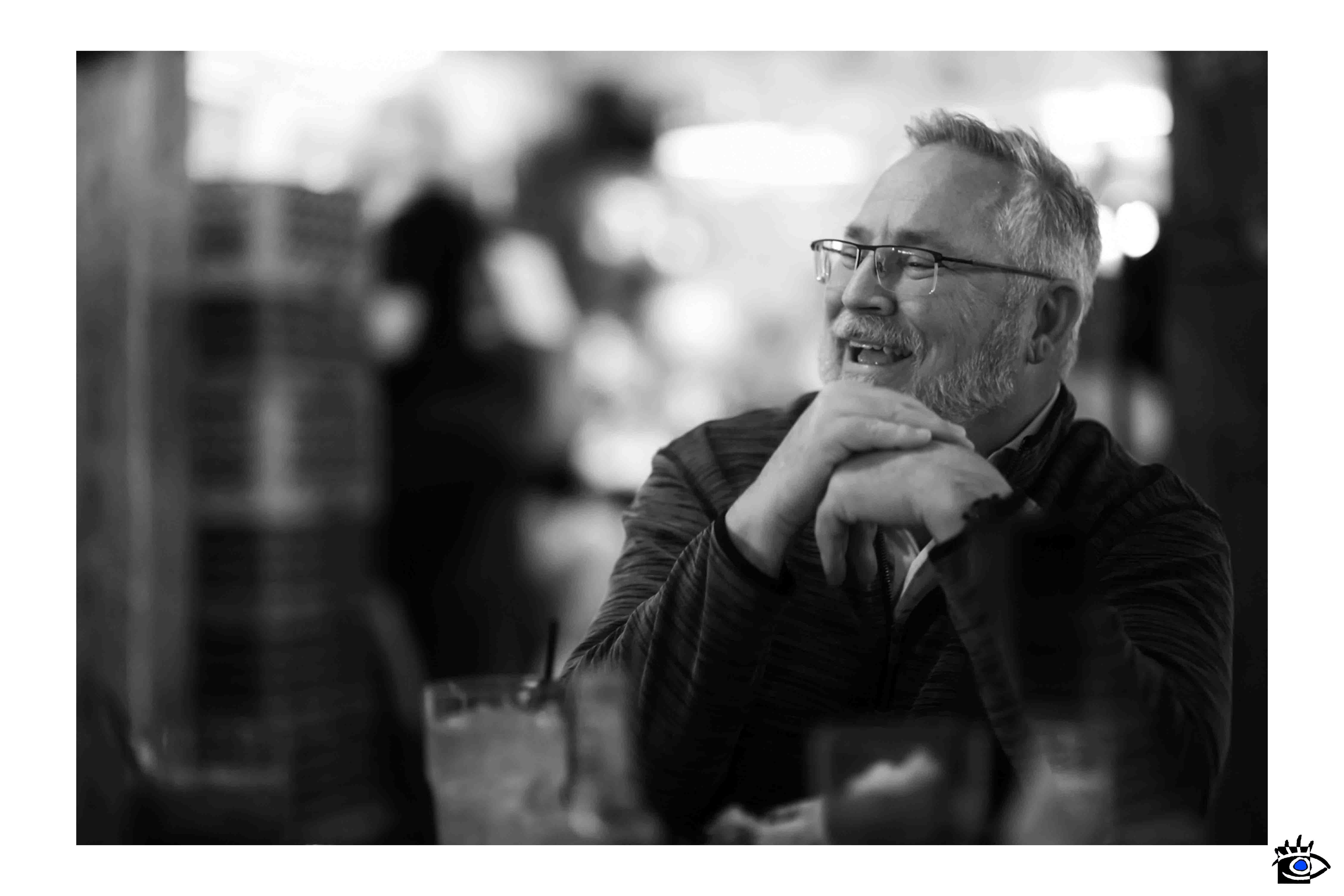 Un uomo anziano ride seduto al tavolino di un bar, catturato in bianco e nero in un momento spontaneo, incarnando l'essenza della fotografia autentica