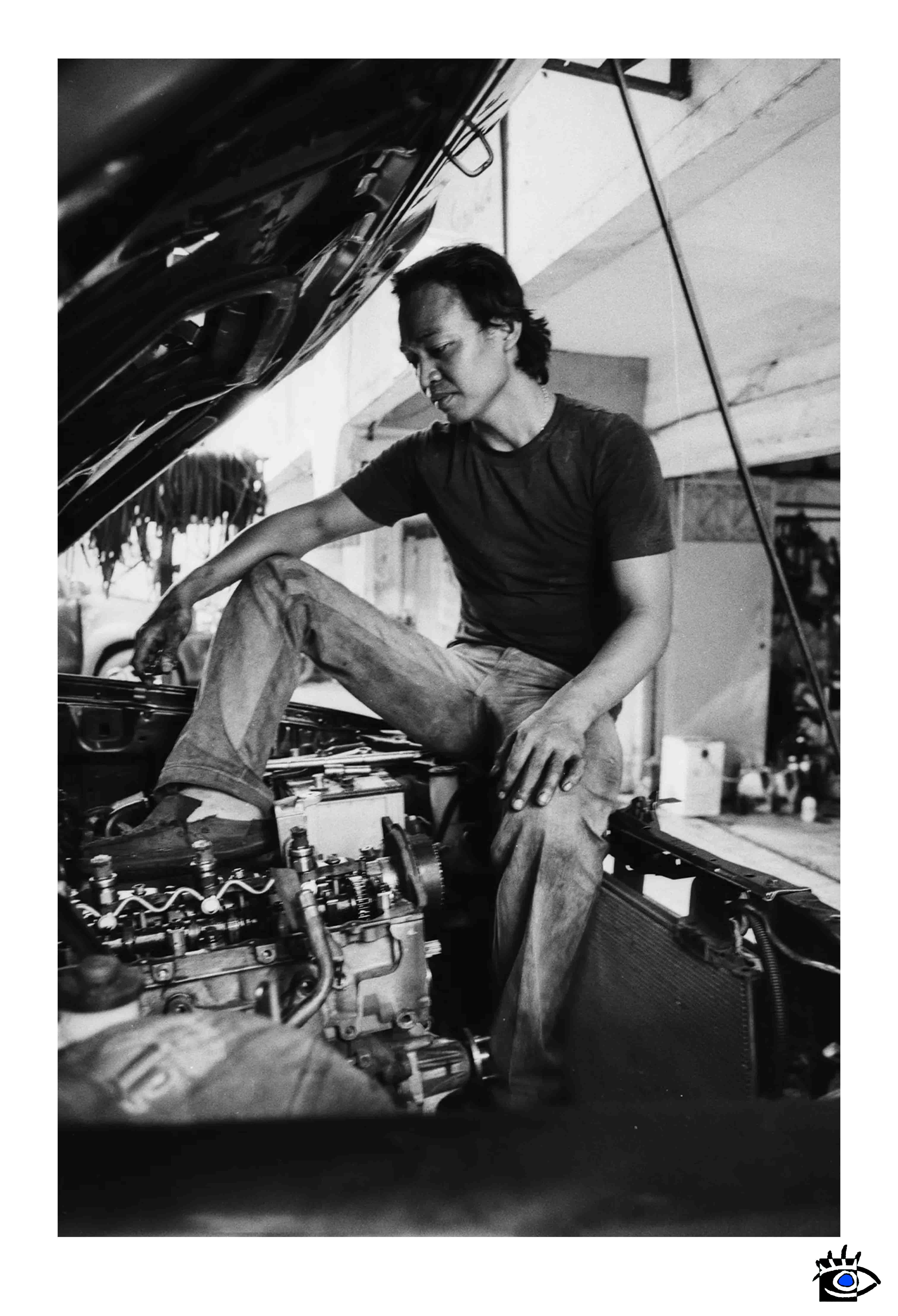 Un uomo assorto nei suoi pensieri, seduto nel vano motore di un’auto americana in un’officina di Natal, Brasile. Una scena autentica catturata durante un viaggio di scoperta e libertà.