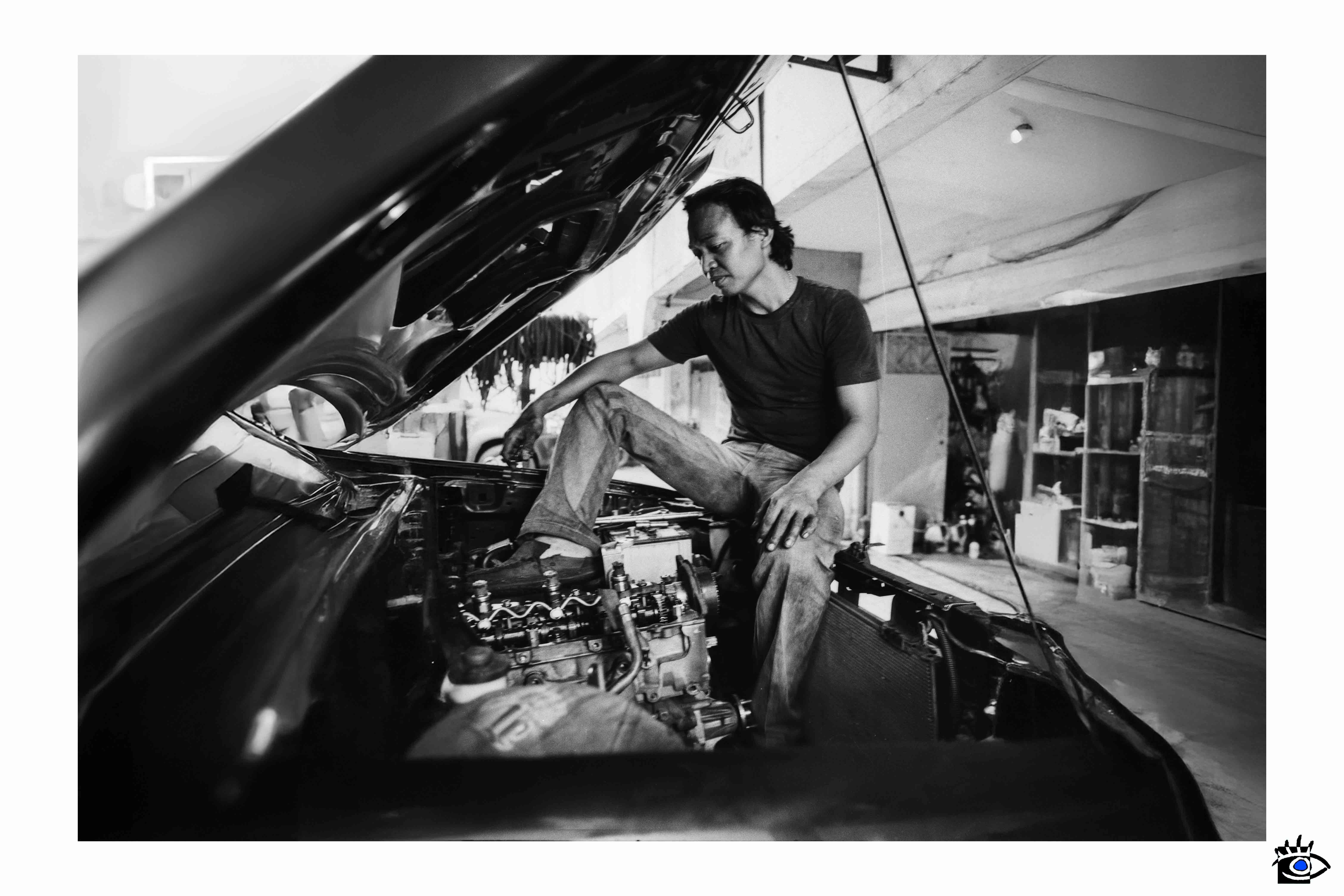 Uomo seduto nel vano motore di una vecchia auto americana in un’officina meccanica, ambientazione grezza e autentica, foto in bianco e nero.