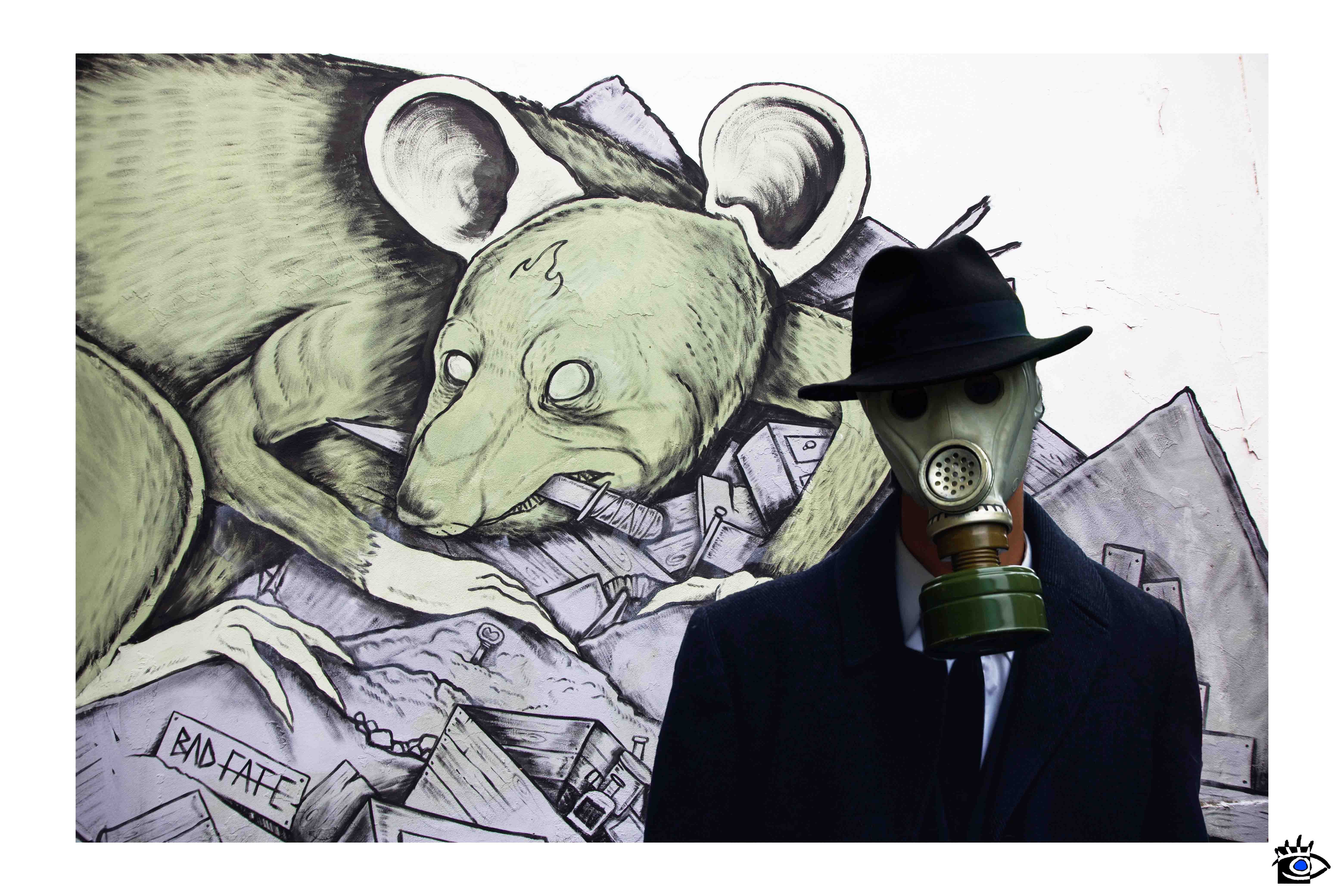 Uomo in cappotto scuro e maschera antigas posa davanti a un murales raffigurante un ratto con occhi vuoti, simbolo di società decadente.