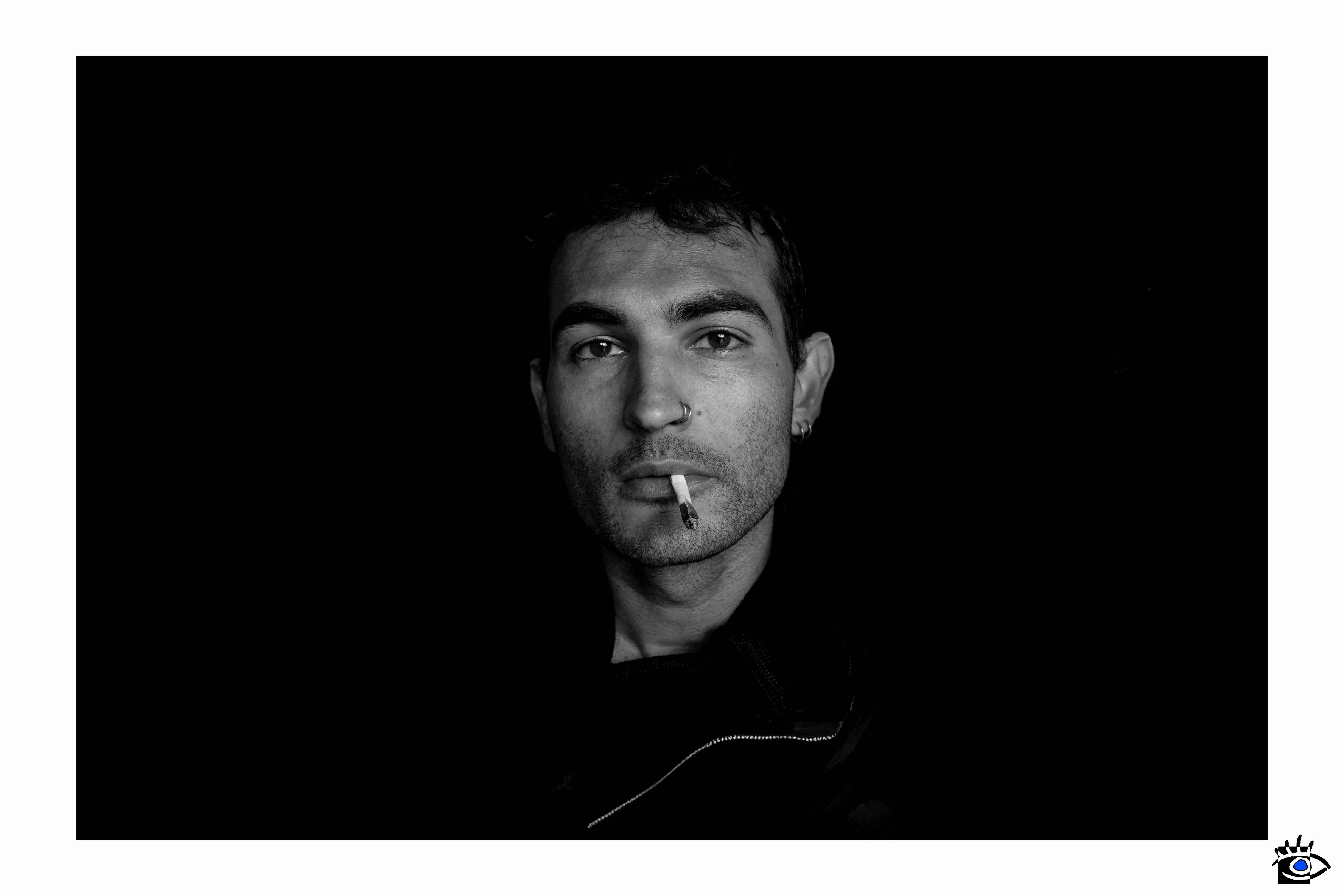 Ritratto in bianco e nero di un uomo con uno spinello spento tra le labbra, uno sguardo diretto e un piercing al naso che riflette una luce tenue.