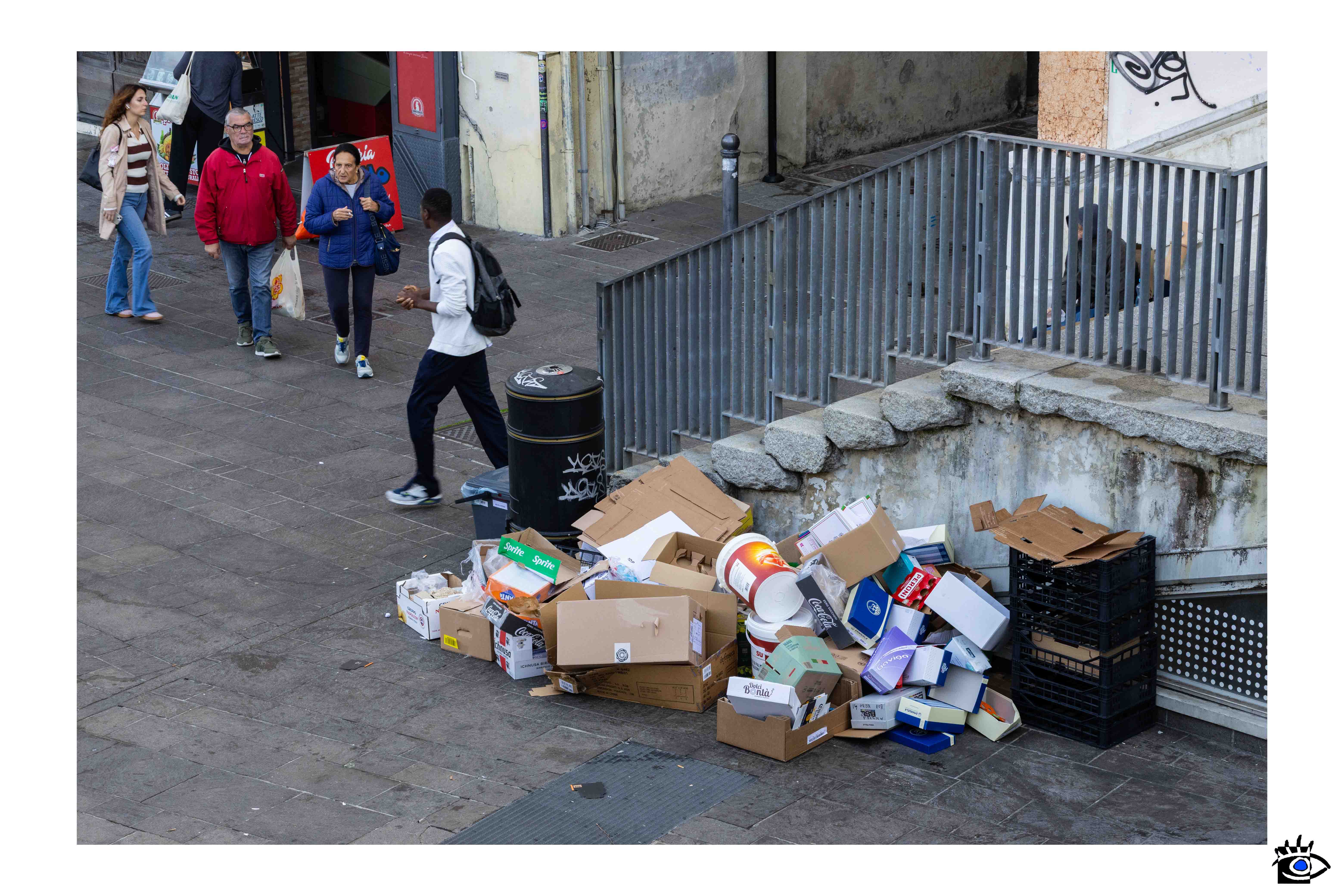 Cumulo di scatole e rifiuti accatastati lungo una strada cittadina, mentre alcuni passanti camminano accanto indifferenti.