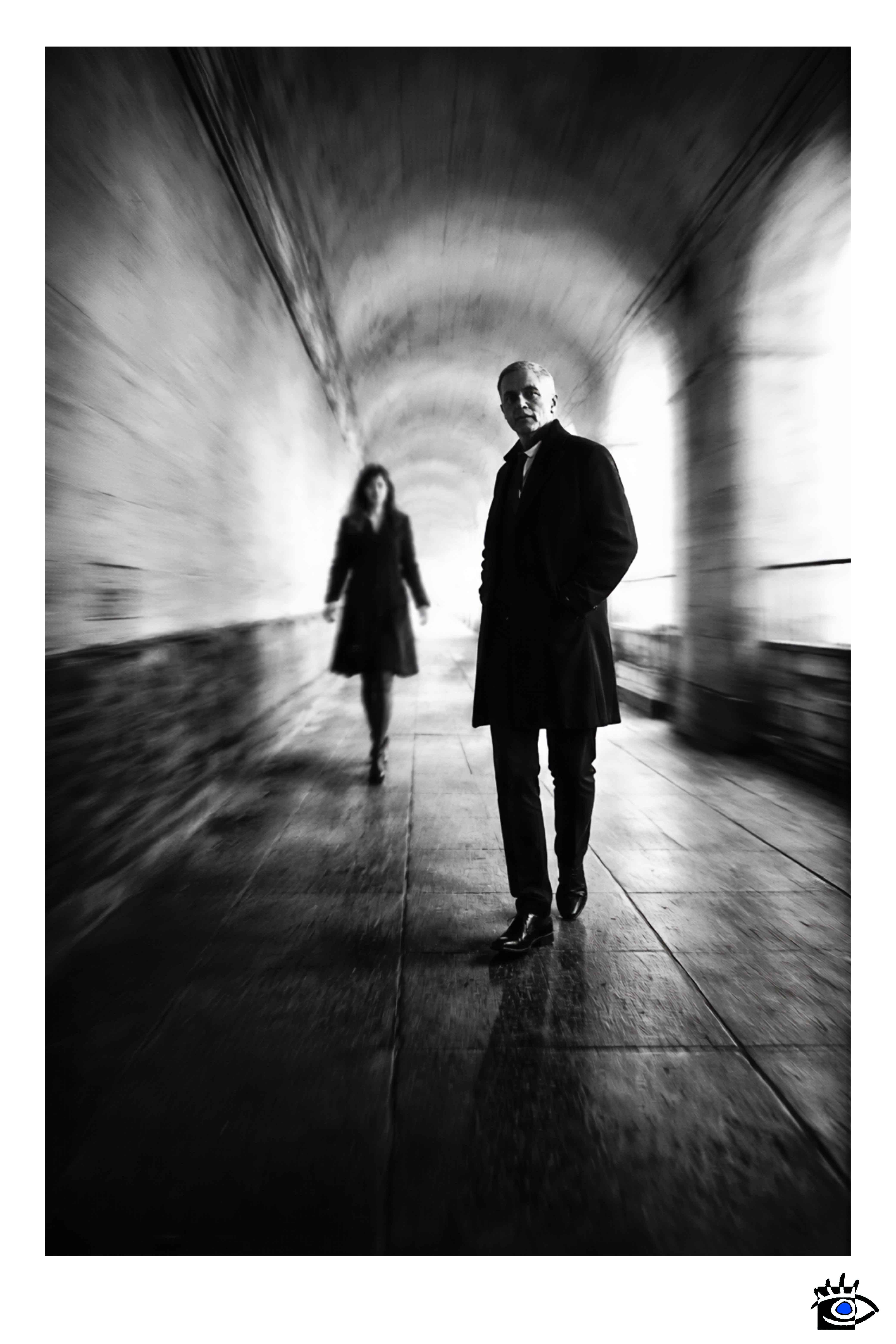 Due figure percorrono una galleria urbana: una in primo piano, l’altra sfocata sullo sfondo, immerse in un movimento che dilata lo spazio e il tempo.