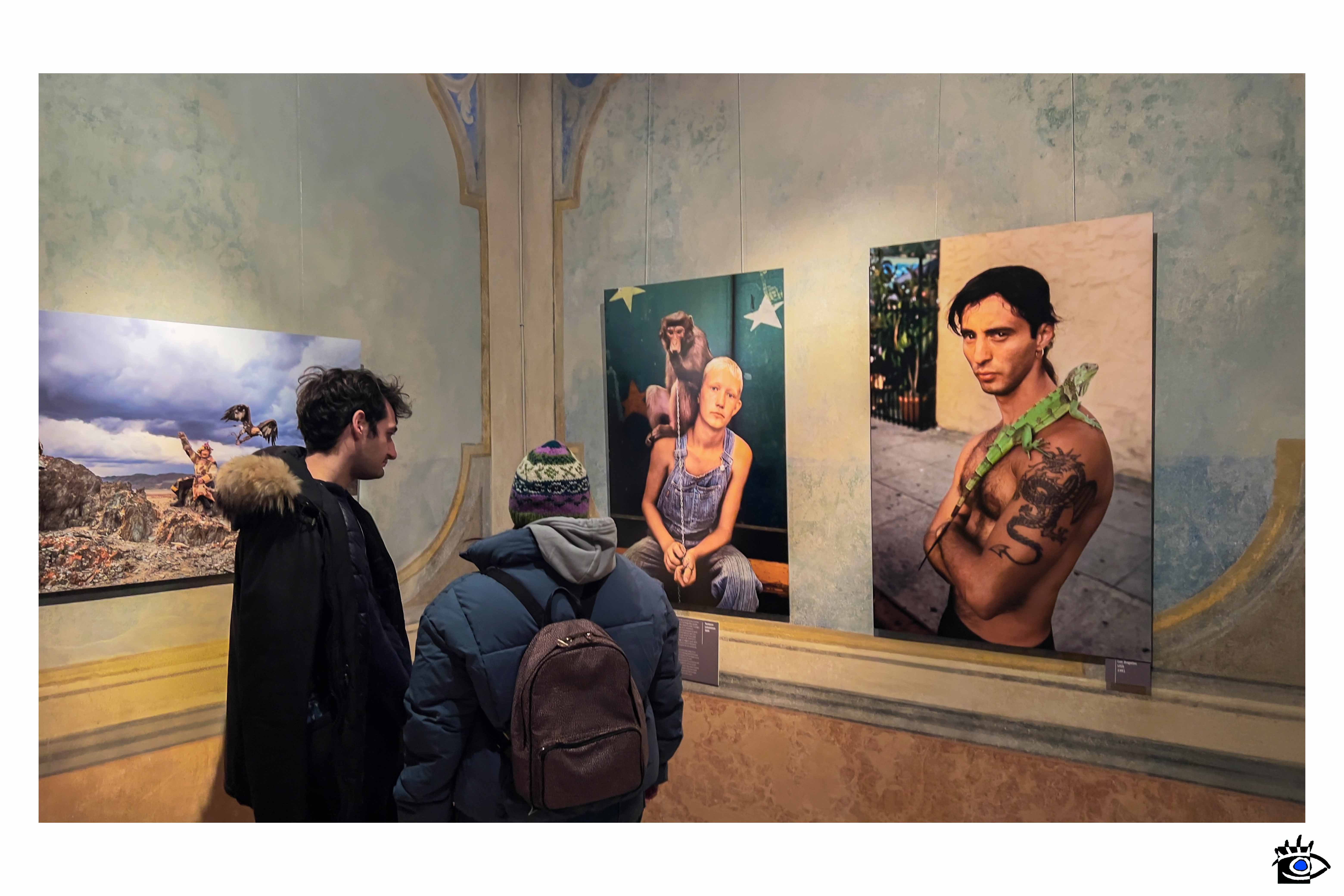 Due visitatori osservano fotografie di grande formato esposte su una parete di una mostra fotografica in un ambiente storico