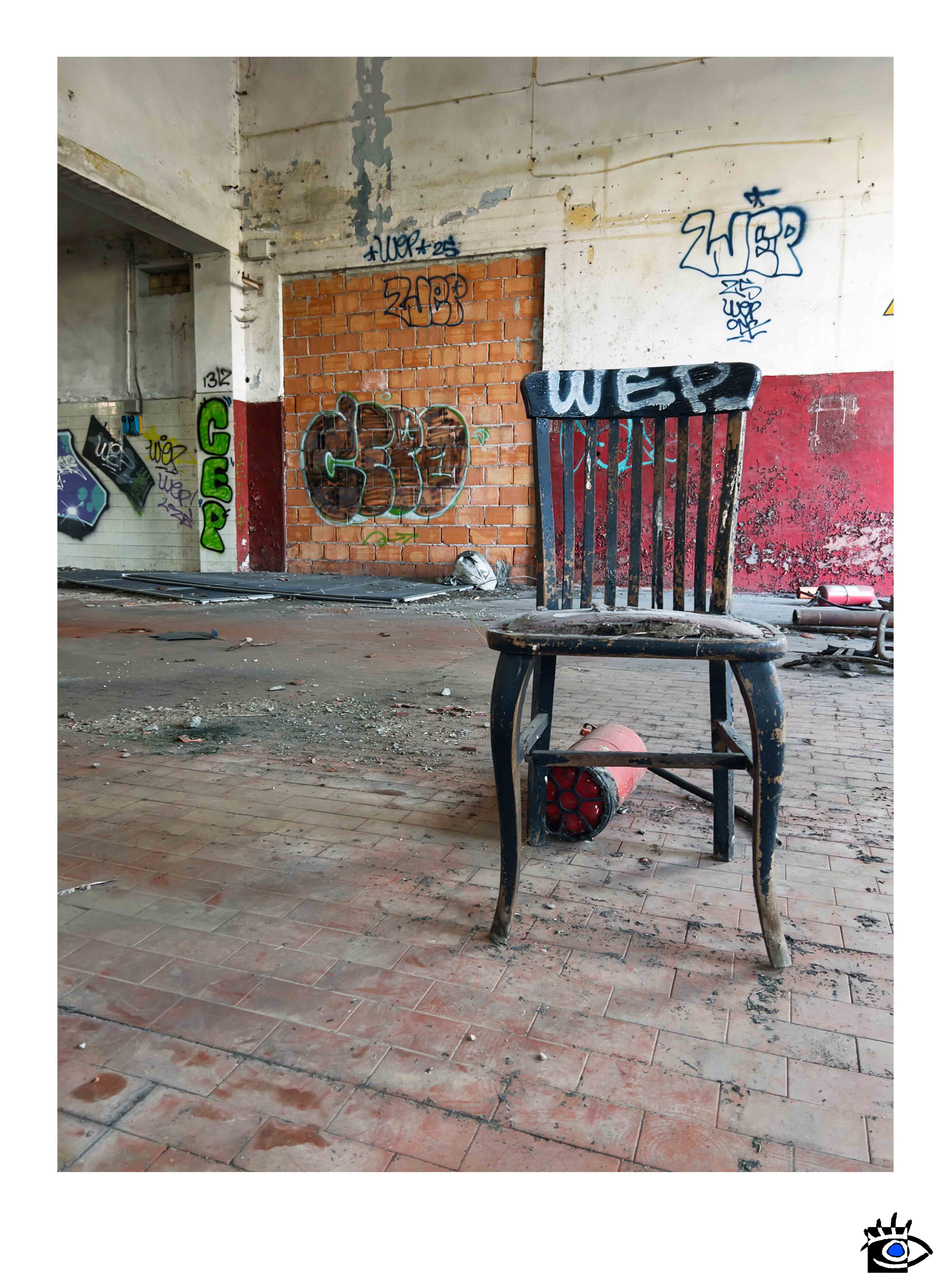 Una sedia in legno rovinata dal tempo è collocata al centro di uno spazio industriale abbandonato, circondata da detriti, graffiti e segni di degrado strutturale.