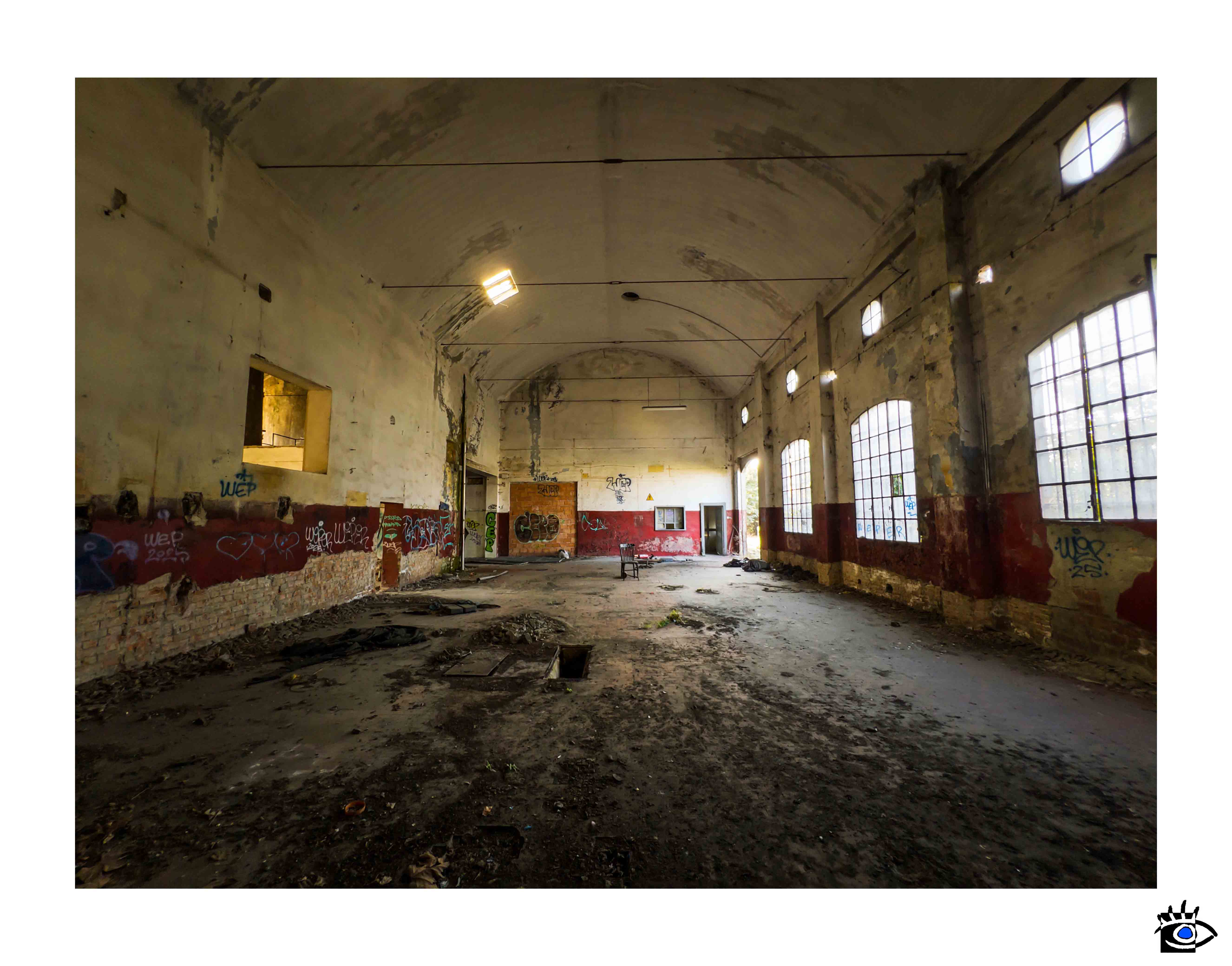 Grande interno di fabbrica abbandonata con pareti scrostate, graffiti, finestre industriali e pavimento coperto di detriti.