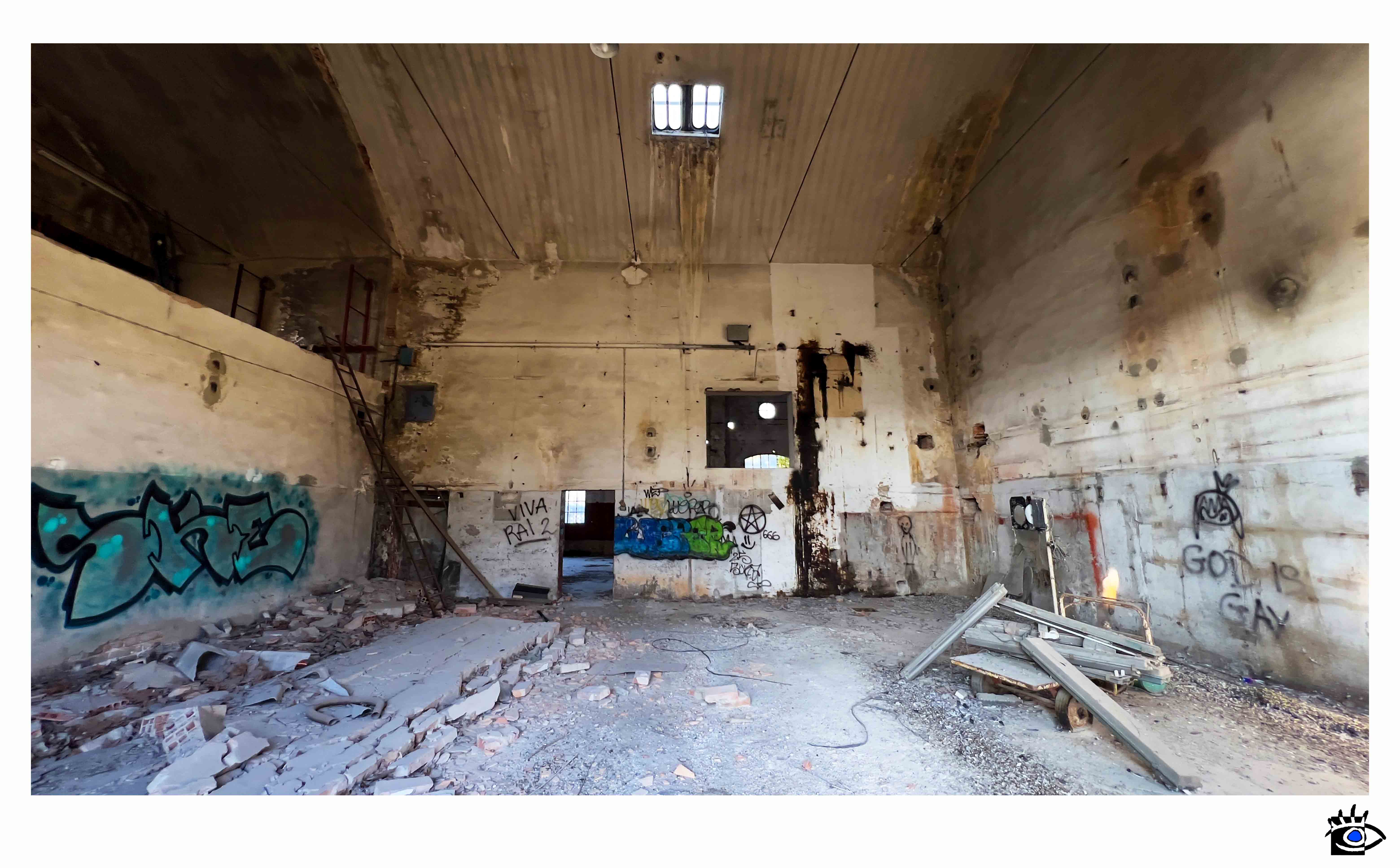 Ampio interno di edificio industriale abbandonato con muri scrostati, graffiti, detriti sul pavimento e luce che entra da finestre alte.