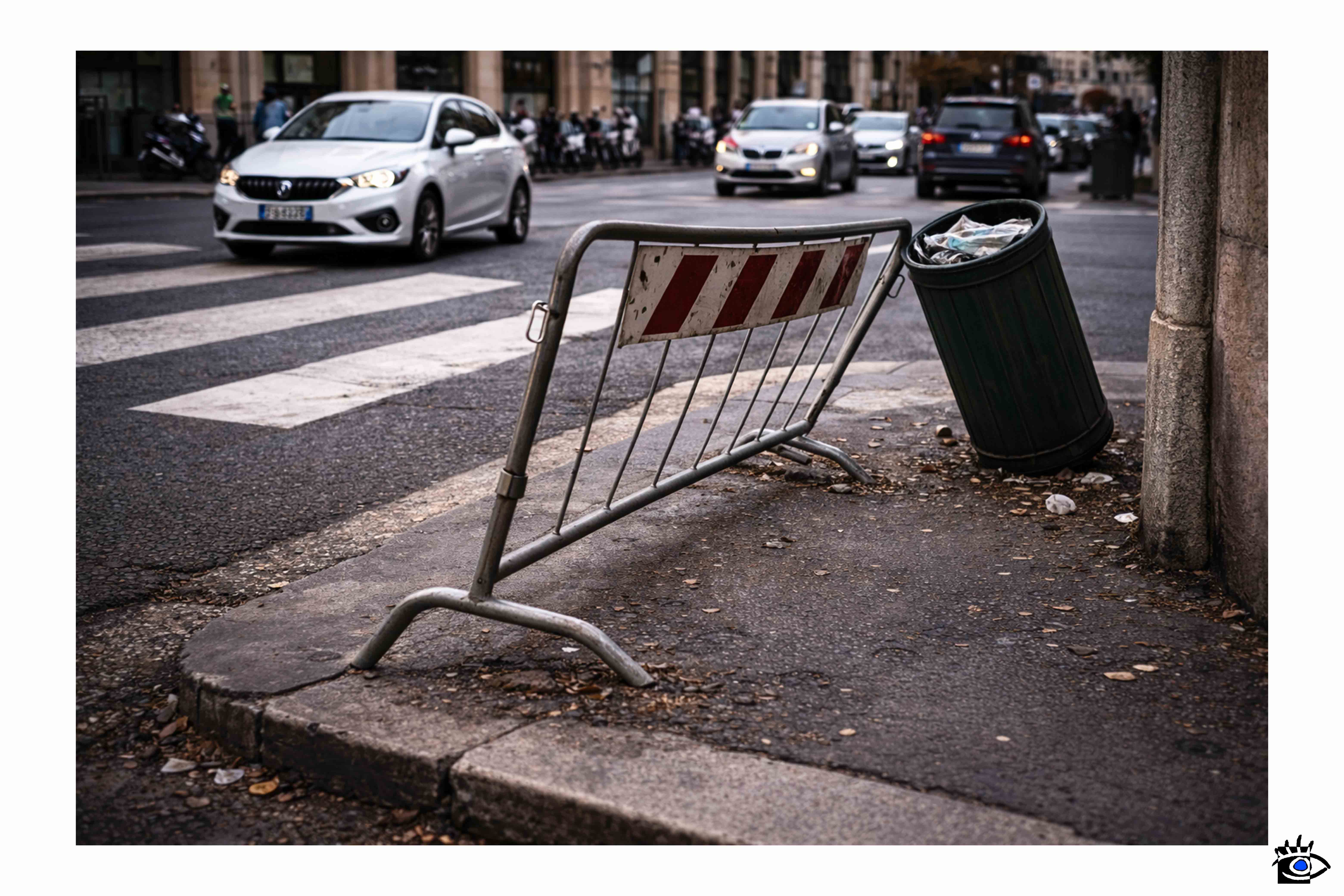 Transenna metallica piegata e cestino spostato su un marciapiede cittadino mostrano come l’uso quotidiano modifichi lo spazio urbano progettato.