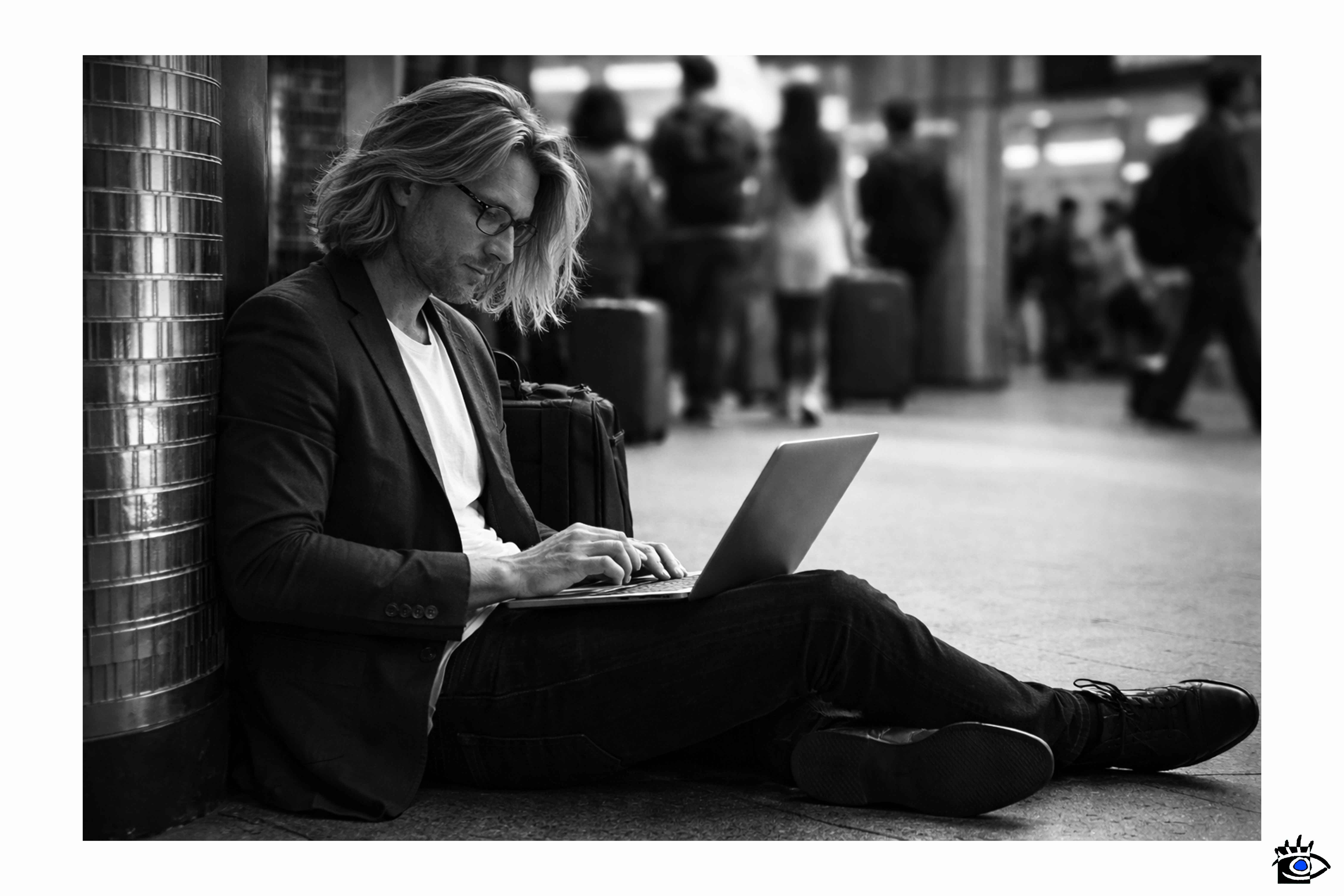 Persona seduta a terra in un luogo di transito usa un computer portatile mentre altre persone passano sullo sfondo con valigie.