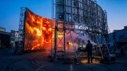 Un enorme schermo LED mostra un volto di intelligenza artificiale in fiamme dall'aria apocalittica, ma la ripresa allargata svela l'impalcatura, i cavi e un tecnico che la produce dalla consolle: l'apocalisse dell'AI come spettacolo costruito.
