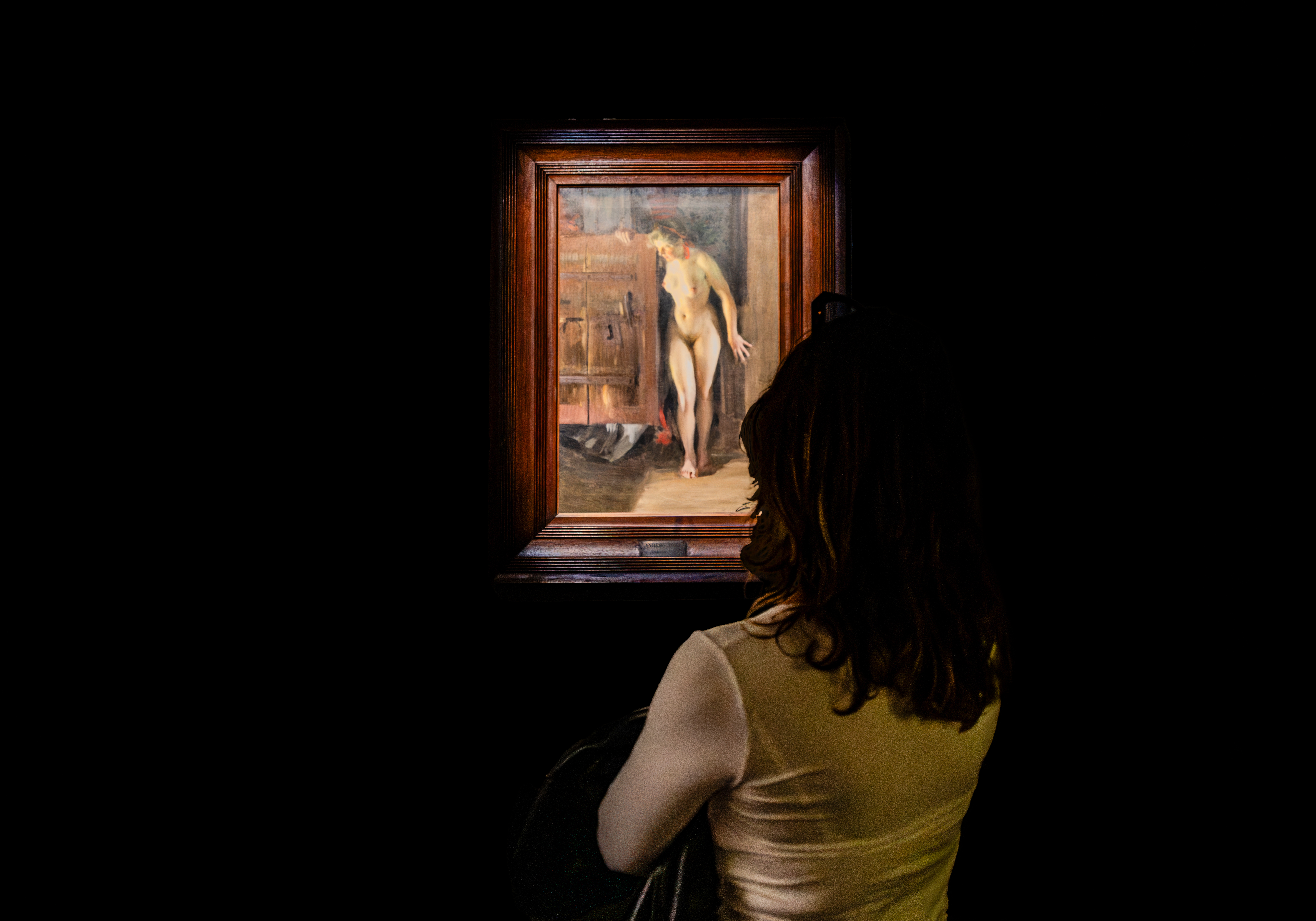 Visitatrice di spalle con capelli ricci e canotta chiara davanti a un dipinto di nudo femminile in una sala buia