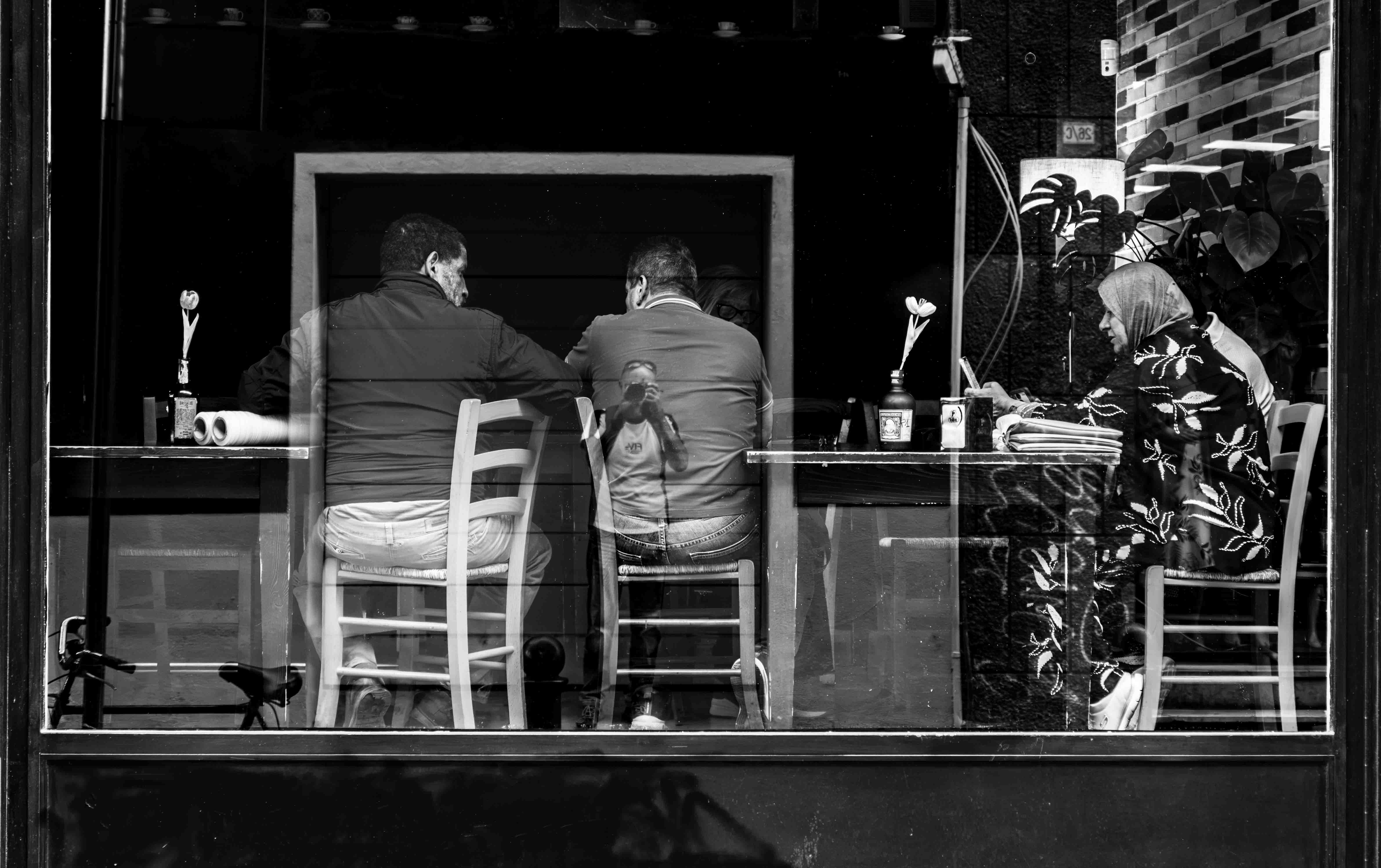 Persone sedute ai tavoli di un bar viste attraverso una vetrina, con il riflesso del fotografo sulla schiena di un uomo